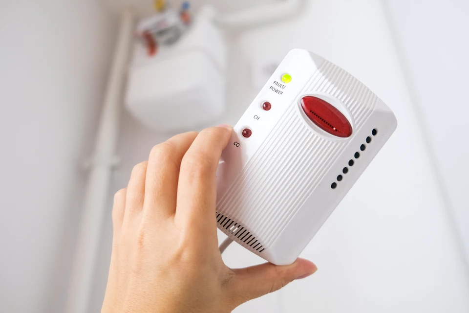 Main dans une maison tenant un détecteur de gaz domestique blanc avec indicateurs LED jaune et rouge, boutons CH4 et CO, grille de ventilation et capteur infrarouge circulaire rouge